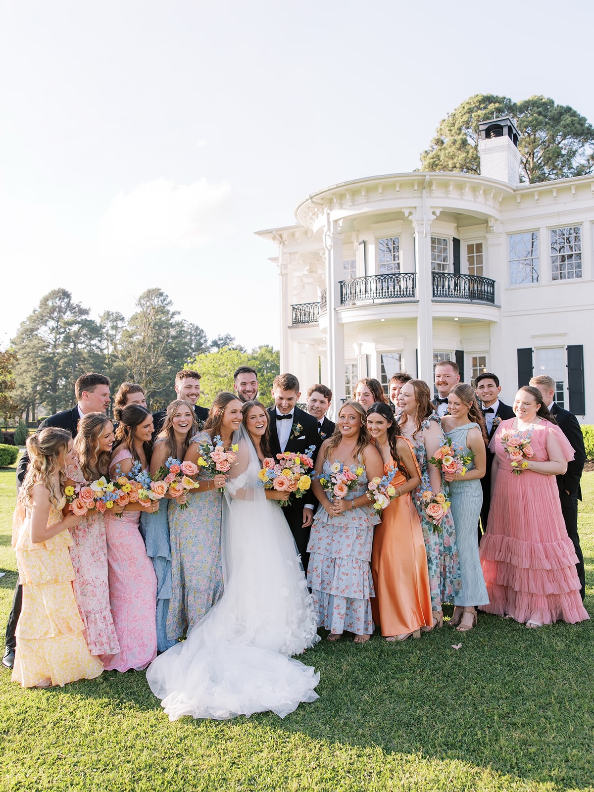 Wedding party photo in front of a white antebellum mansion with bridesmaids in mismatched pastel dresses and groomsmen in black tuxedos
