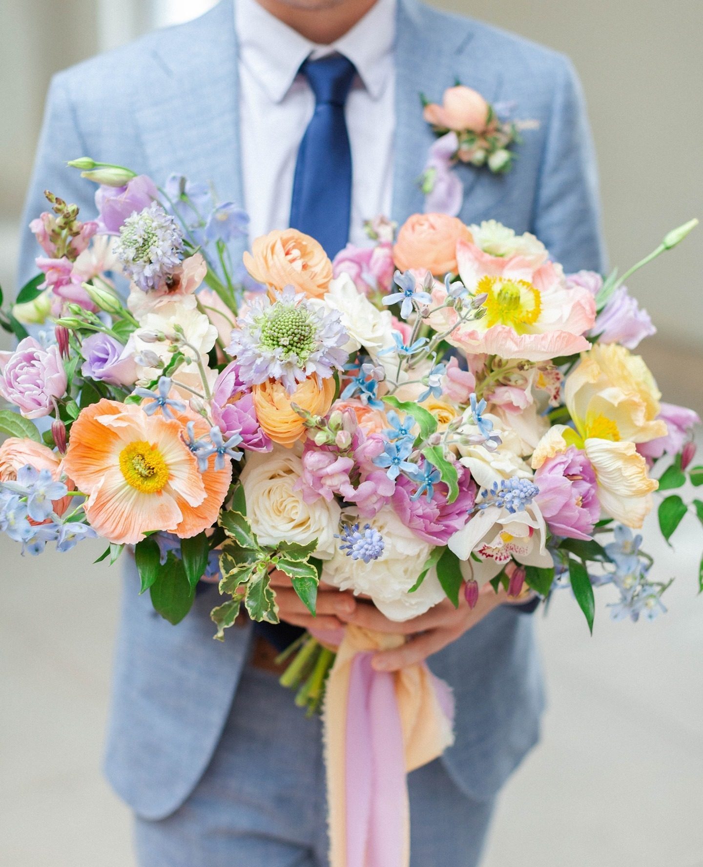Groom in a light blue suit and navy tie holding a lush multi-pastel bridal bouquet of peach, lavender, blush, blue, and yellow florals