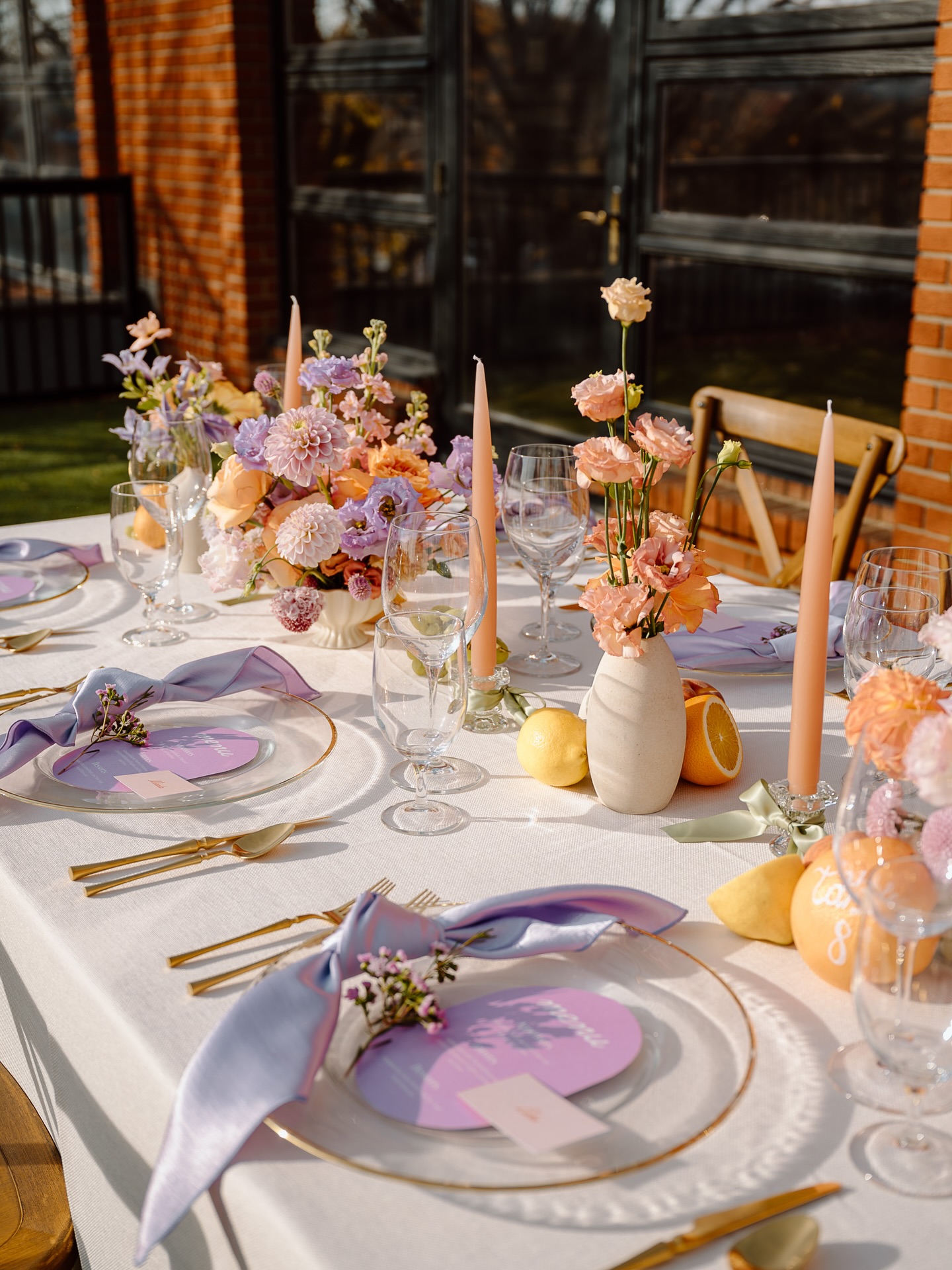 Outdoor pastel wedding tablescape with peach taper candles, peach and lavender florals, gold flatware, and citrus styled on a linen runner