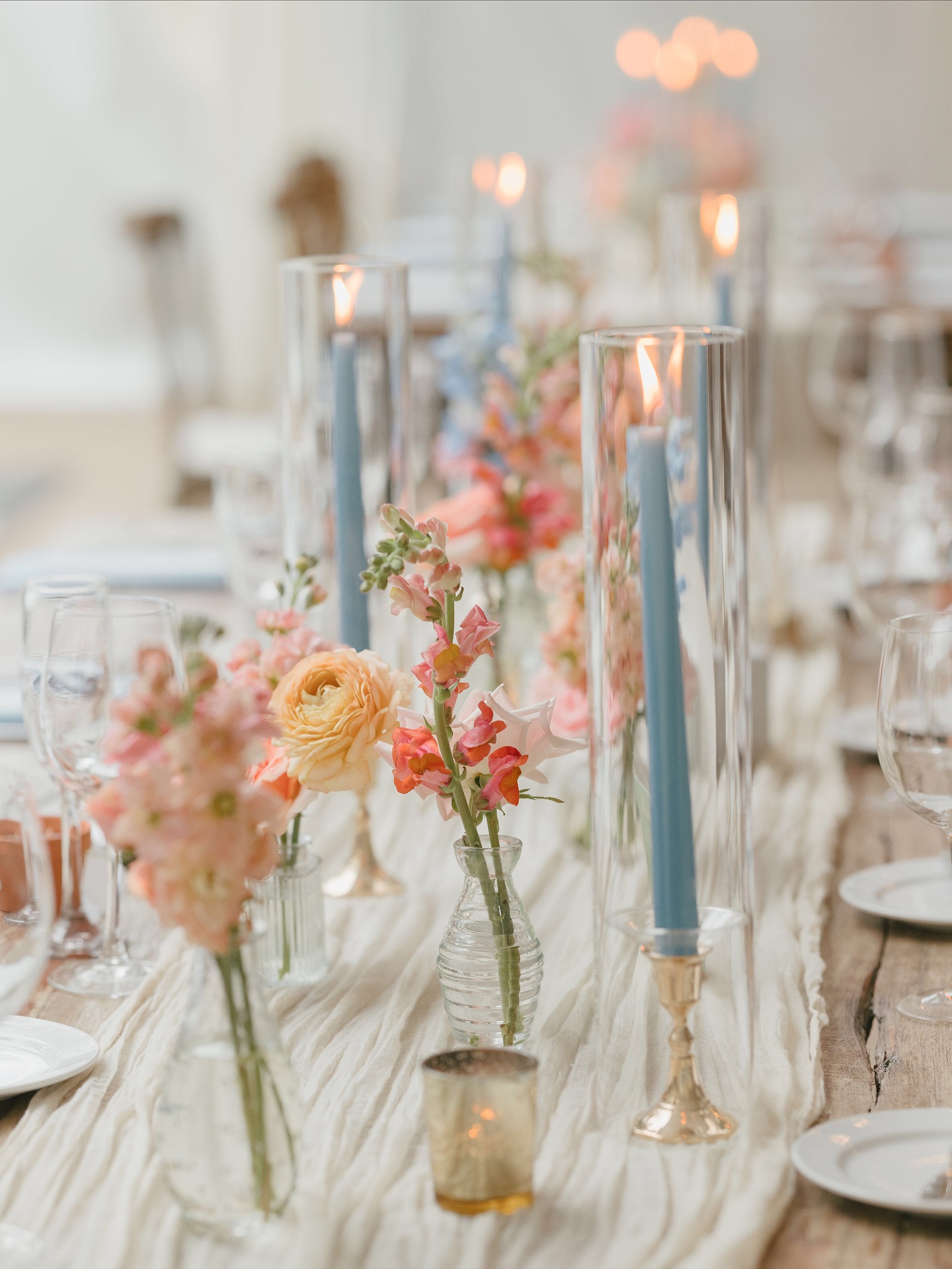 Pastel wedding reception tablescape with peach and pink bud-vase florals, dusty blue taper candles in tall hurricanes, and brass candlesticks on a cheesecloth runner