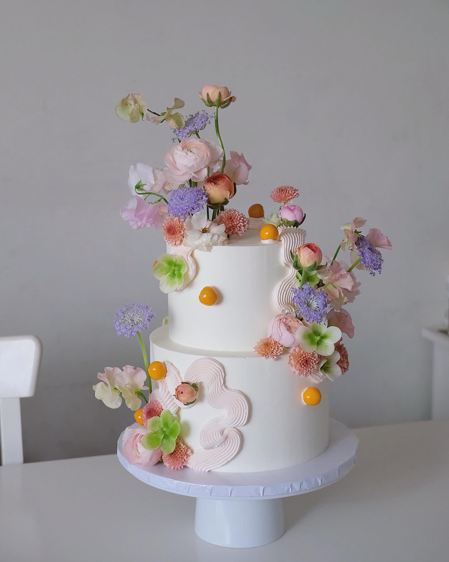 Two-tier white buttercream wedding cake decorated with peach, blush, lavender, and sage hellebore florals and yellow pearl accents