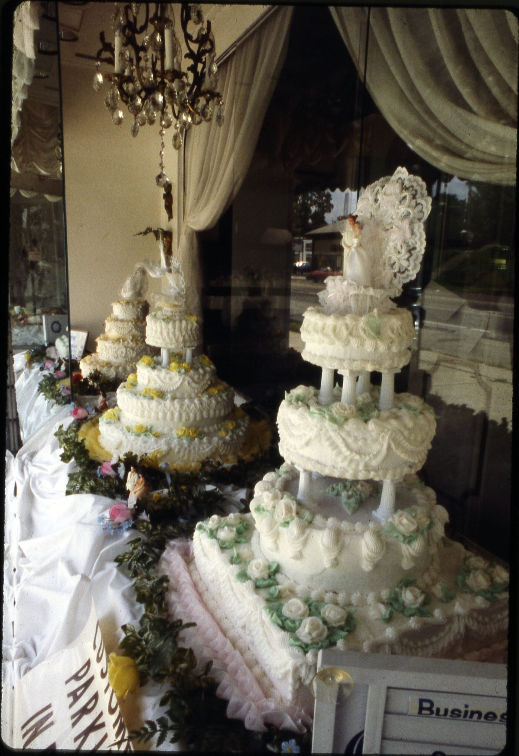 Vintage bakery display window with three multi-tier wedding cakes on white pillars, decorated with ruffled buttercream, garlands, and lace toppers, under a chandelier