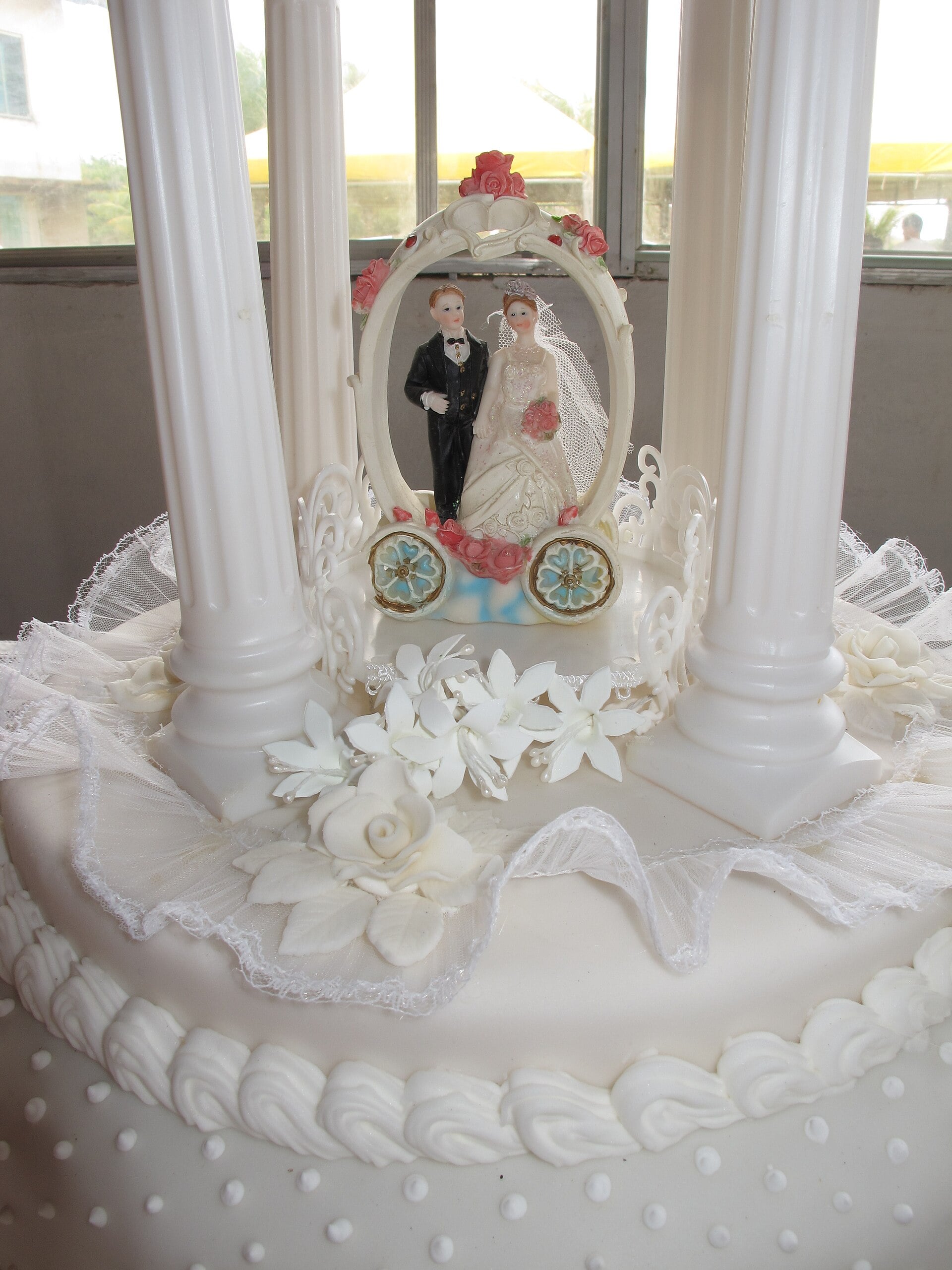 Wedding cake topper of a bride and groom framed inside a white oval arch, set between white columns on a ruffled cake with lace and pearl details