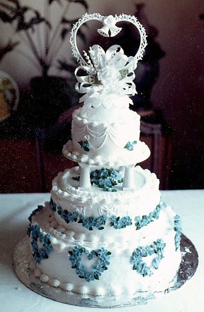 Three-tier 1980s style wedding cake with white frosting, blue piped borders and blue flower accents, topped with a heart-shaped frame