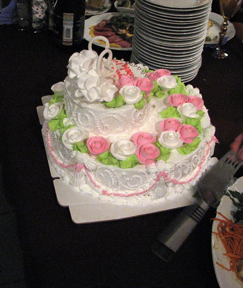 White wedding cake covered in ruffled royal icing borders, pink drapes, green leaves, and piped buttercream roses