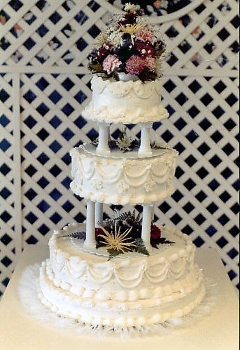 Three-tier wedding cake separated by white Grecian pillars with shell borders and cascading icing swags, photographed against white lattice