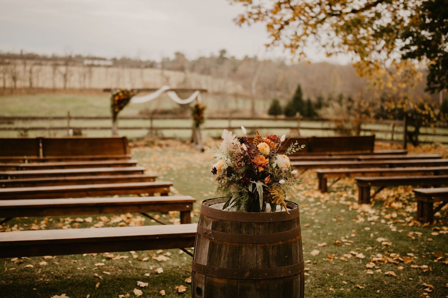 Rustic fall wedding ceremony arrangement of orange and burgundy flowers on a wine barrel between wooden benches