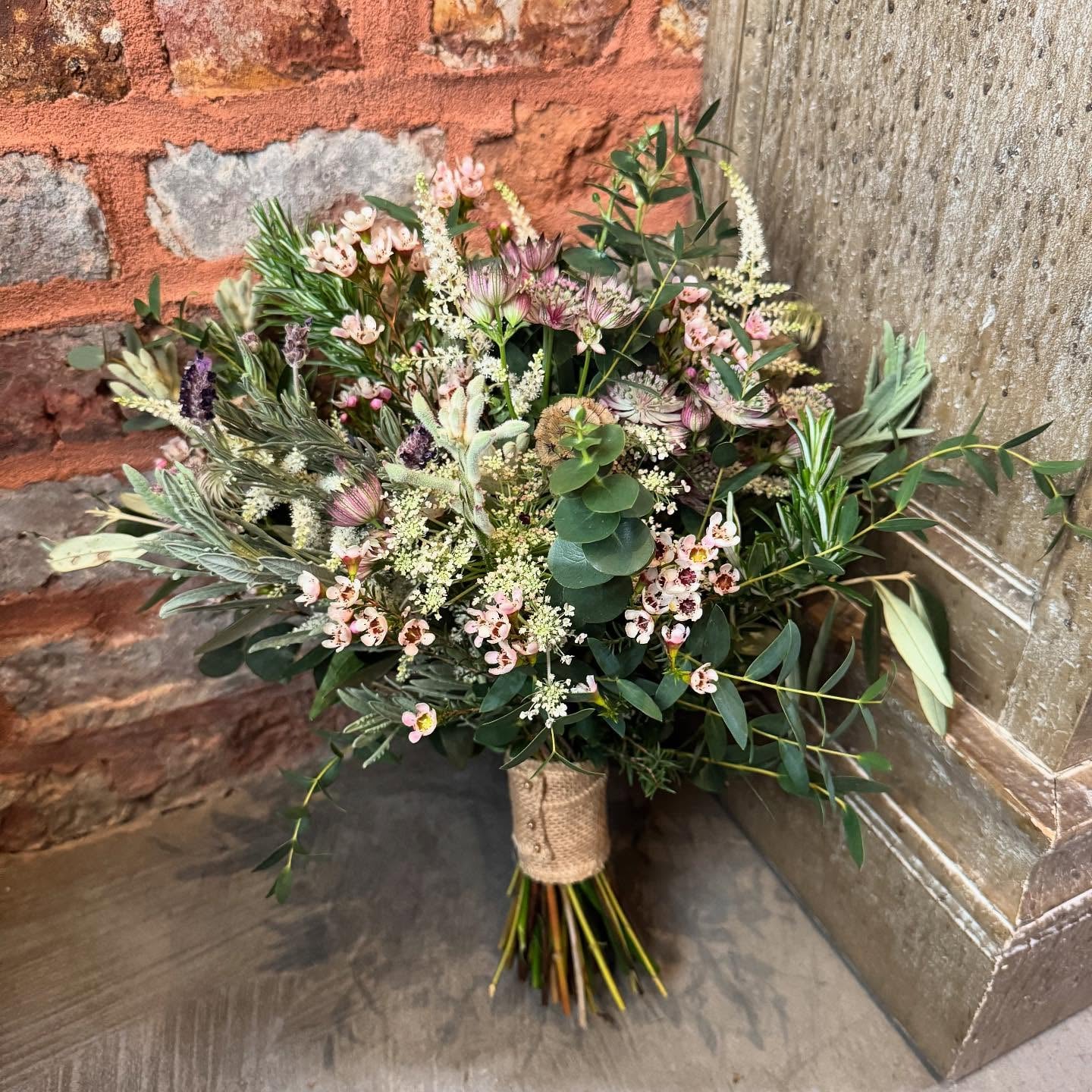 Rustic woodland bridal bouquet with waxflower, lavender, herbs and mixed greenery wrapped in burlap against a brick wall
