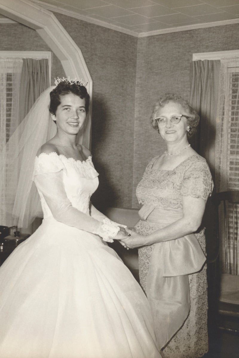1950s bride in a white gown and veil holding hands with her mother