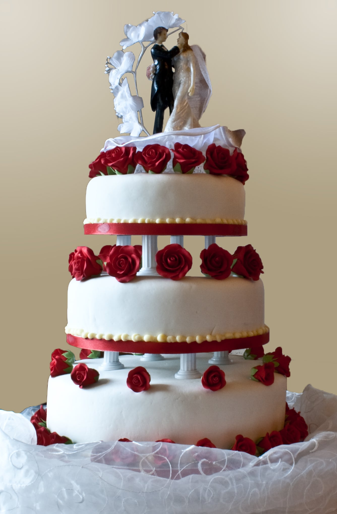 Tiered wedding cake on short pillars with red roses between the tiers and a bride and groom topper under a white arch