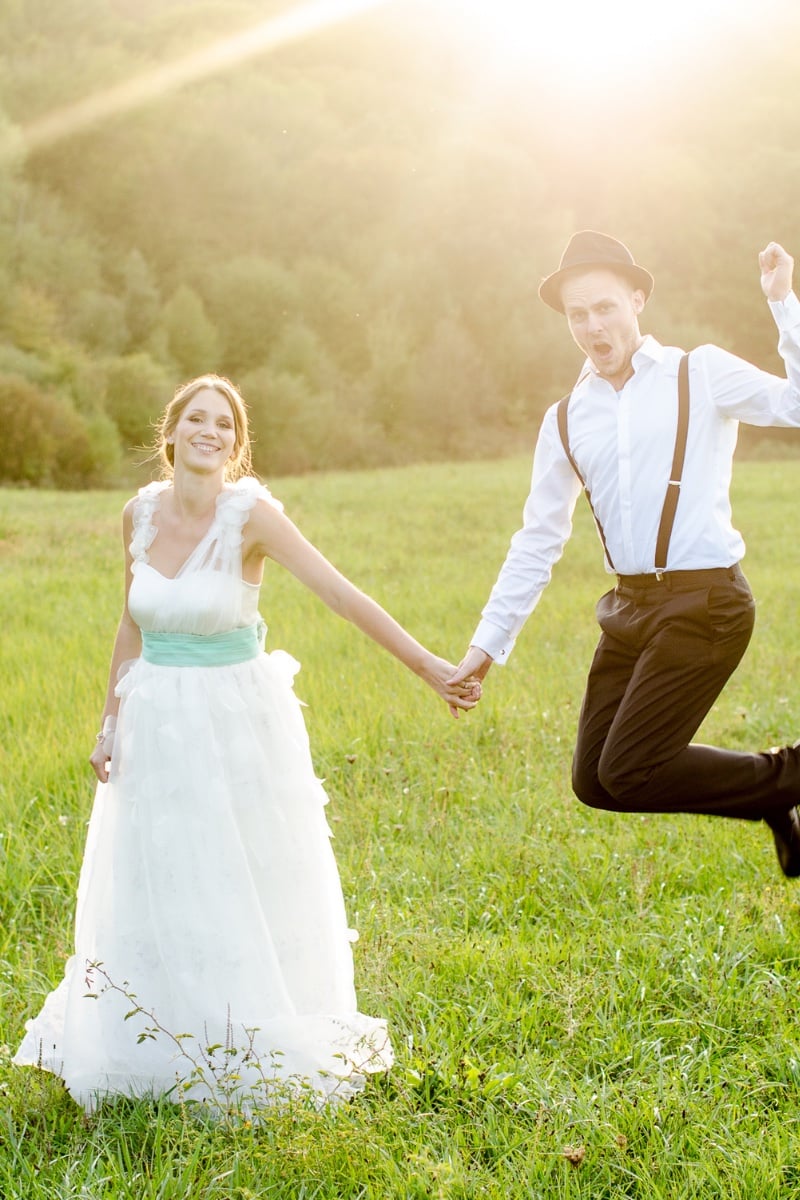 Bride and groom holding hands in a field at golden hour while the groom jumps for joy