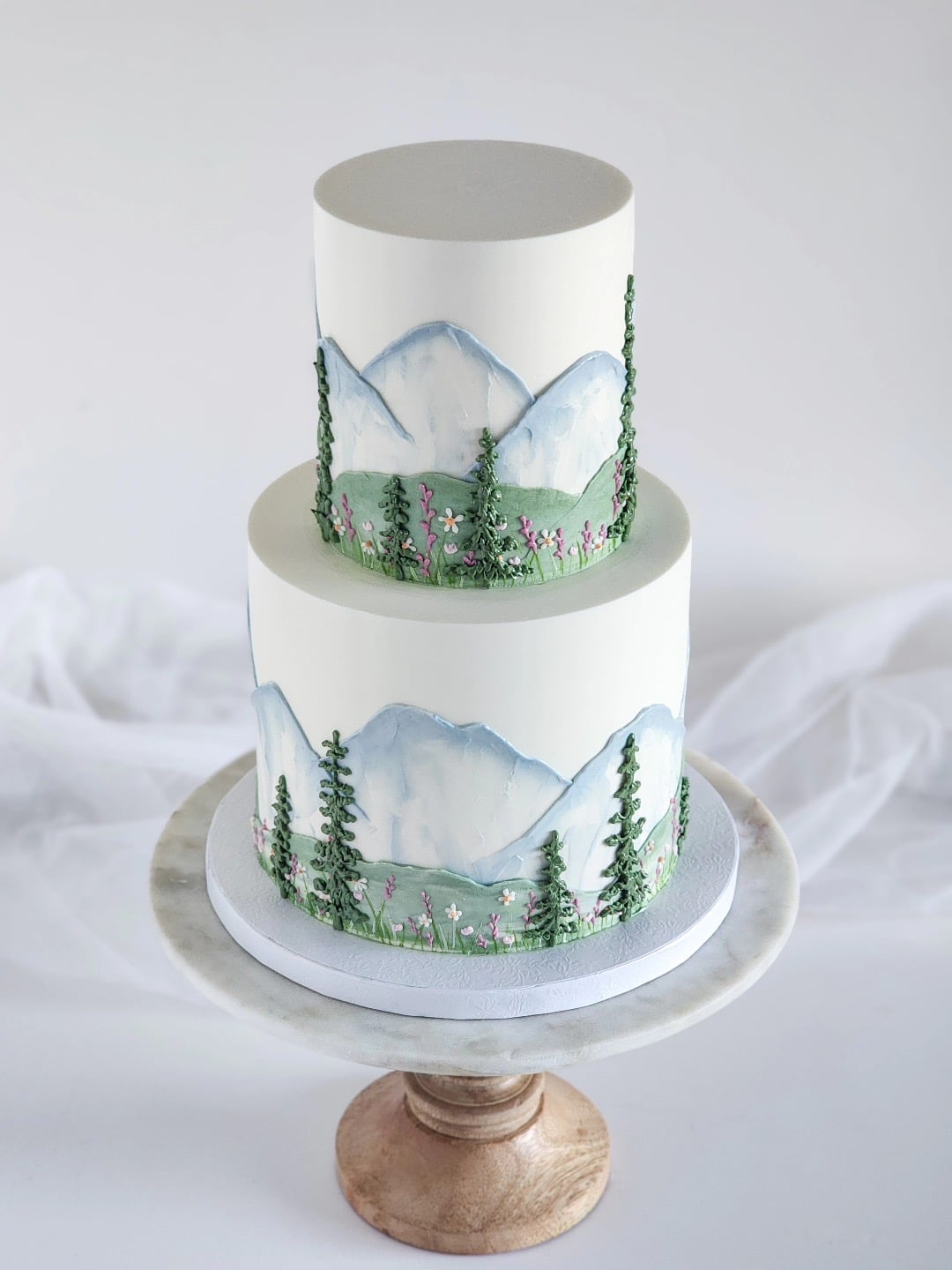 White two-tier wedding cake painted with watercolor mountains, evergreens, and wildflowers