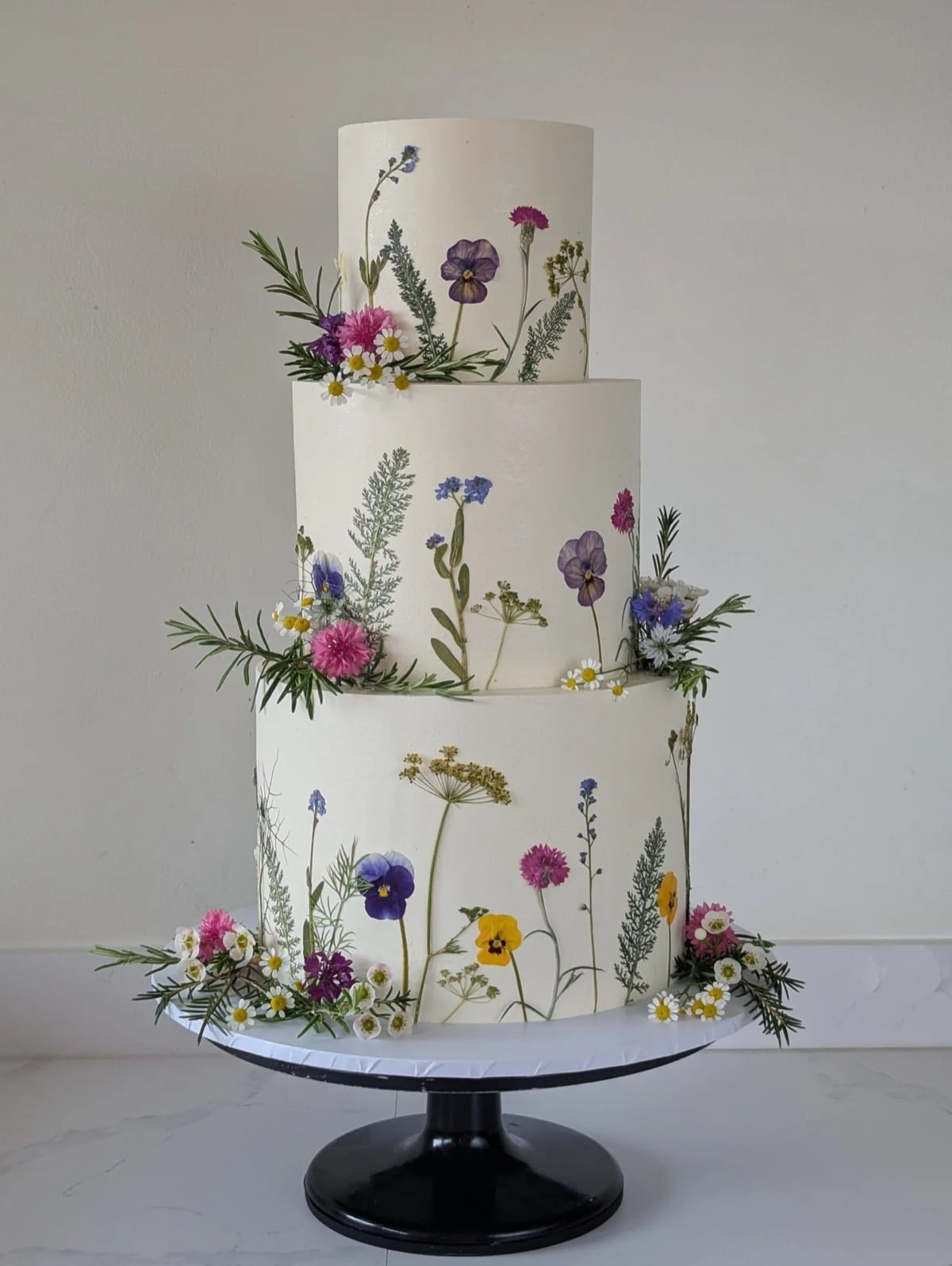 Three-tier white wedding cake decorated with pressed edible wildflowers including pansies and chamomile
