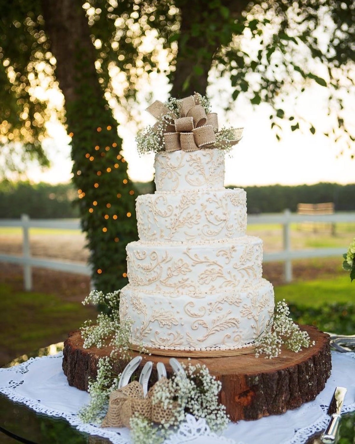Ivory wedding cake with scrollwork piping, a burlap bow topper, and baby's breath on a tree trunk slice