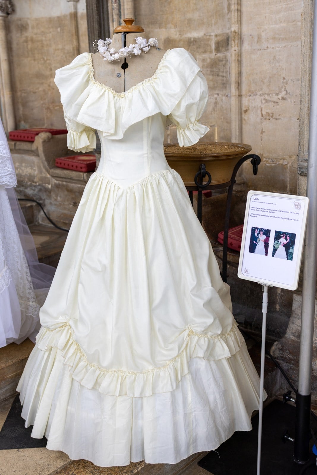 1987 Pronuptia wedding dress on a mannequin with a ruffled off-the-shoulder collar, huge puffed sleeves, drop waist, and a ruffled tiered skirt with bows, at the Ely Cathedral bridal gown exhibition