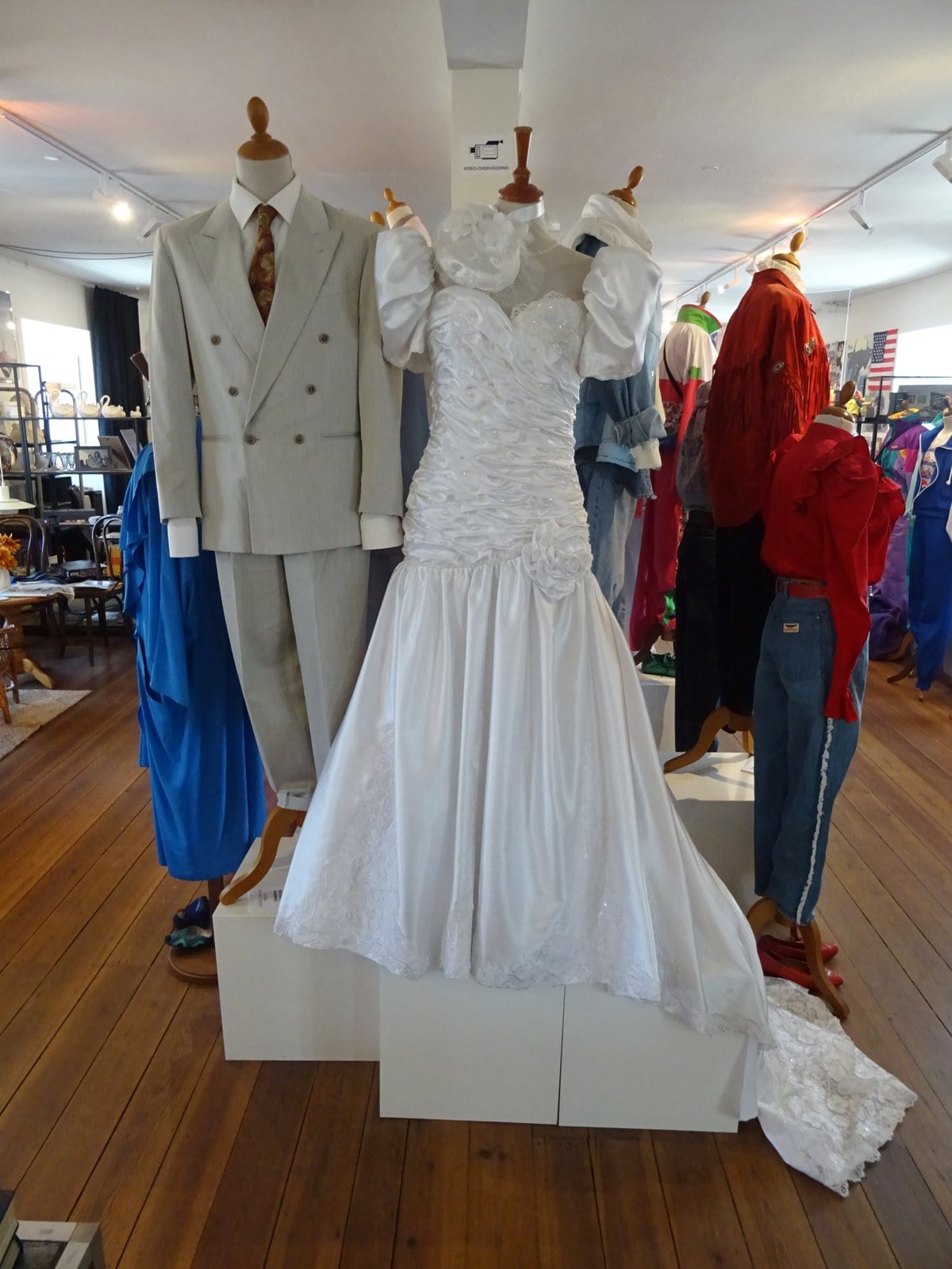 1980s ruched satin drop-waist wedding dress on a mannequin next to a groom's grey double-breasted suit at the Ung i 80'erne exhibition in Odense, Denmark