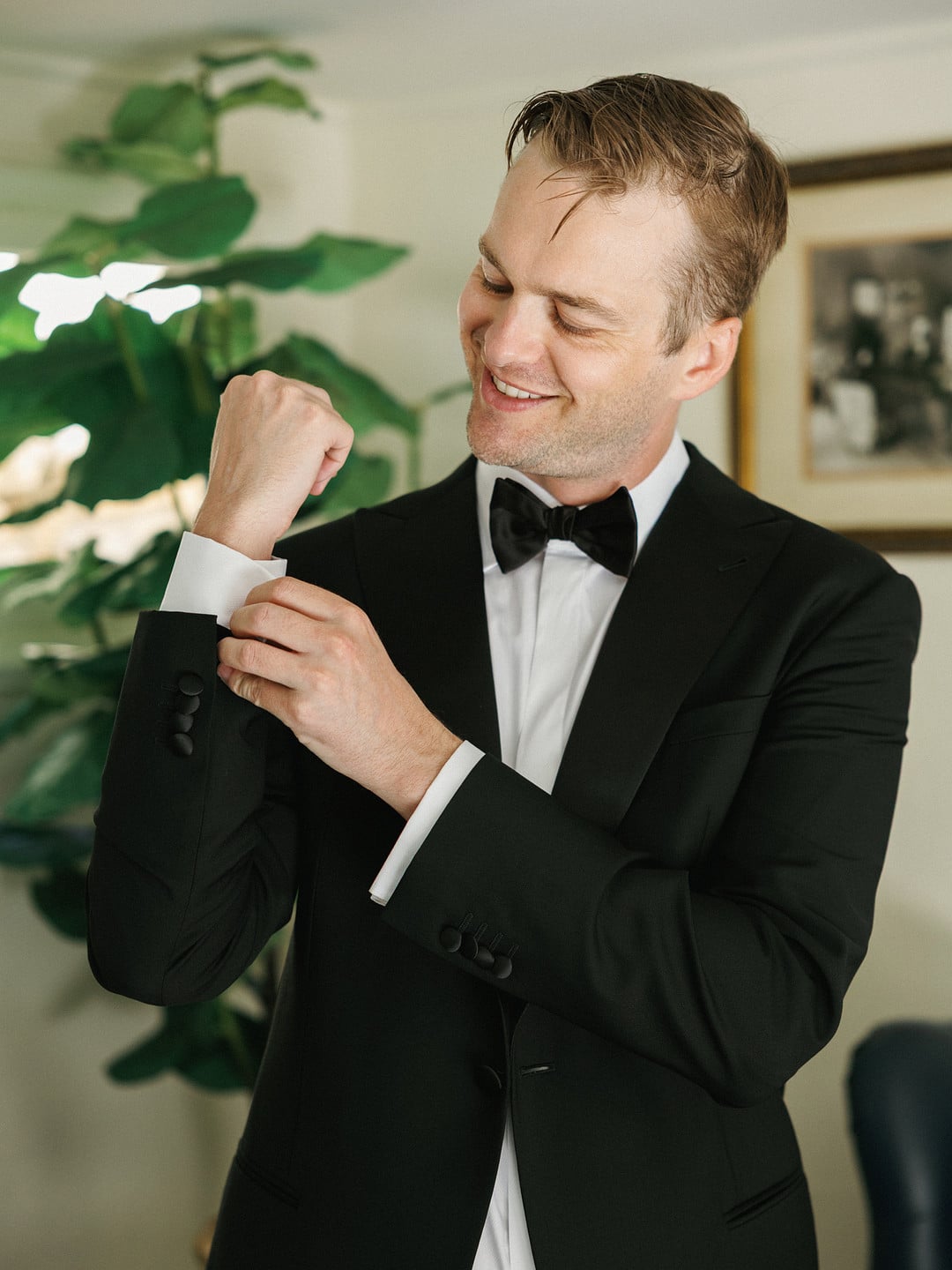 Avery smiling while fastening his cufflinks in a black tuxedo