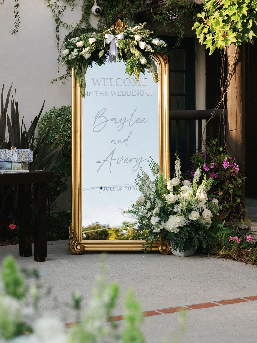 Gold mirror welcome sign surrounded by white and green florals at the reception