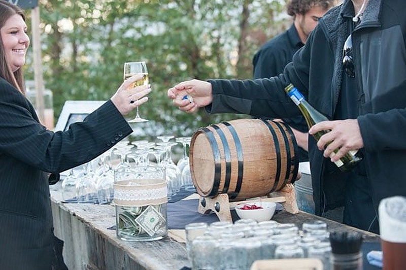 Guests being served wine from a small barrel at an outdoor wedding bar