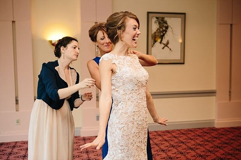 Bride in a lace gown laughing while her mother in navy and a friend help her get ready