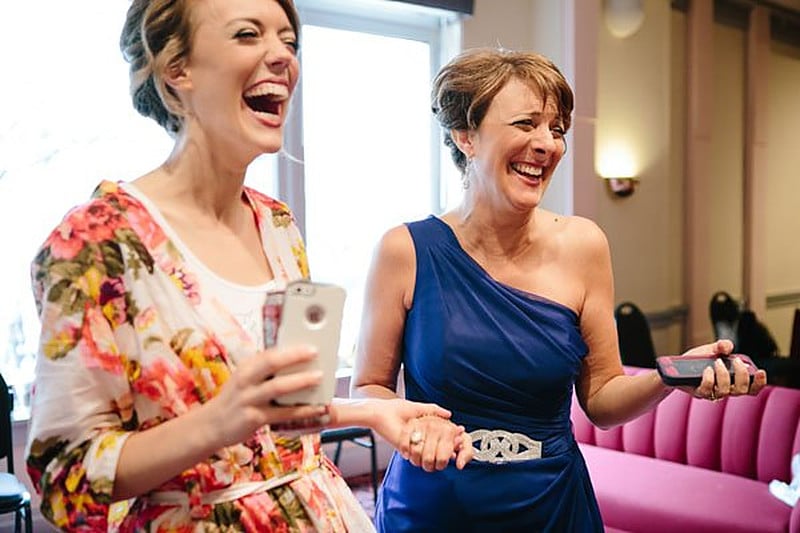 Bride in a floral robe and her mother in a blue dress laughing over their phones before the wedding