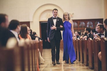 Groom walking down a church aisle arm in arm with his mother in a purple gown