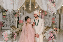 Riya in a blush pink lehenga walking toward Jay at the white carved mandap