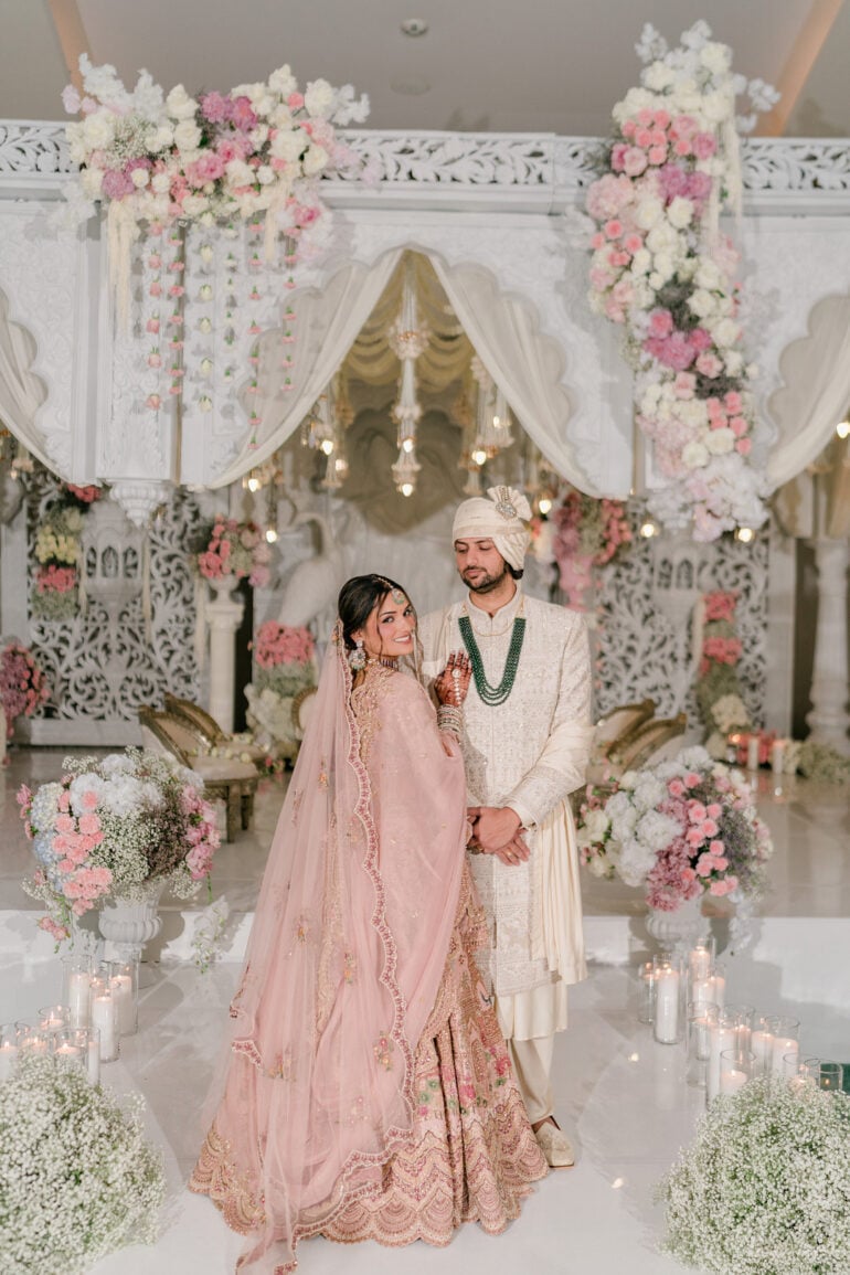 Riya in a blush pink lehenga walking toward Jay at the white carved mandap