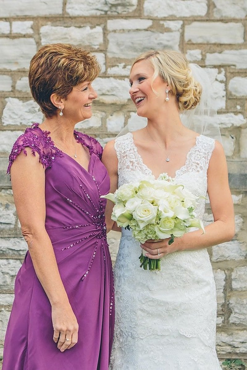 The mother of the bride in a purple lace dress laughing with the bride outside a stone church
