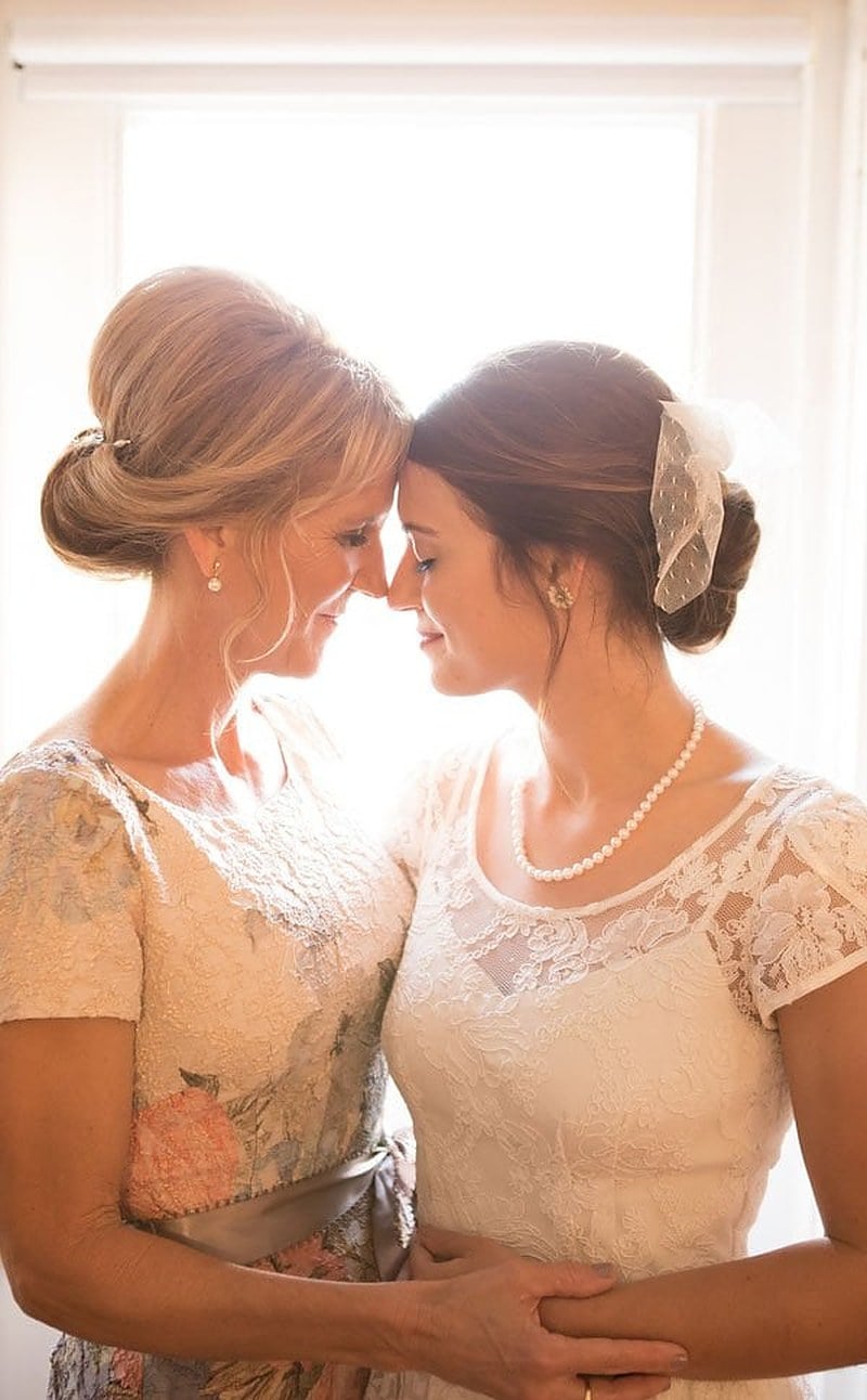 A mother and her daughter, the bride, forehead to forehead in front of a bright window