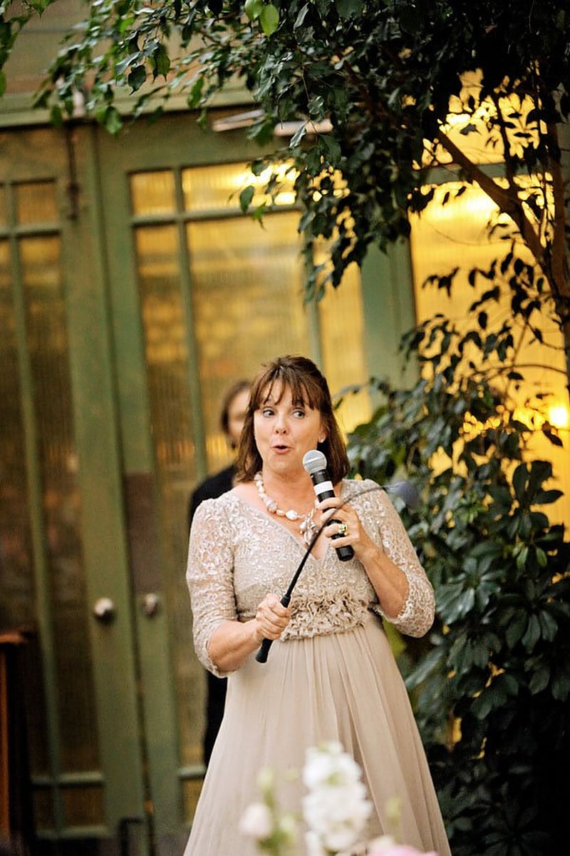 Mother in a champagne lace dress giving a toast into a microphone at an outdoor reception