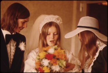1974 bride holding a large round bouquet of orange, yellow, and white daisy mums with the groom and a bridesmaid in a wide-brimmed hat