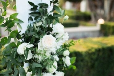 Wedding ceremony arch column wrapped in greenery and white roses with white draping