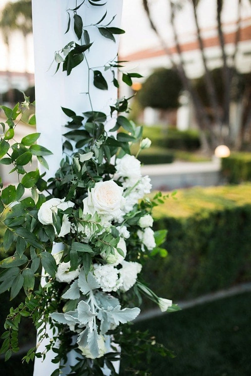 Wedding ceremony arch column wrapped in greenery and white roses with white draping