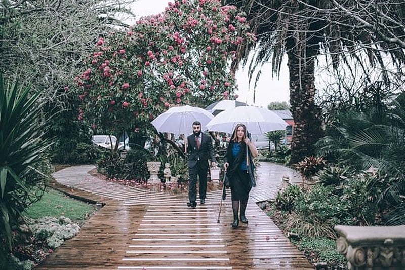 Wedding guests walking a rain-soaked garden path under white umbrellas at Markovina Vineyard Estate in New Zealand