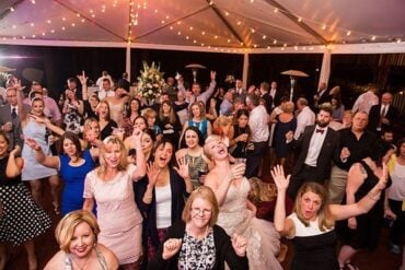 The bride laughing with wedding guests who have their hands in the air on a string-lit reception tent dance floor